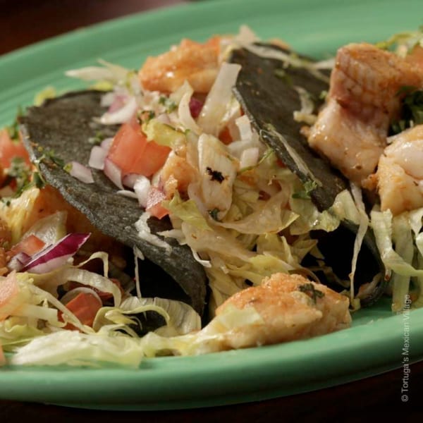 Close-up of three shrimp tacos with blue corn tortillas, lettuce, tomato, and onion on a green plate.