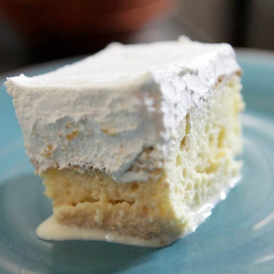 A slice of Tres Leches cake with white frosting and milk pooling on a blue plate.