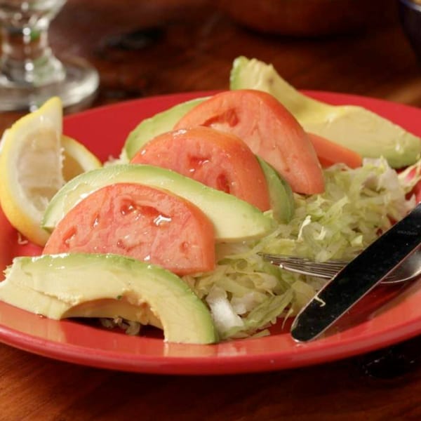 A red plate of shredded lettuce topped with sliced tomatoes, avocado, and lemon wedges.
