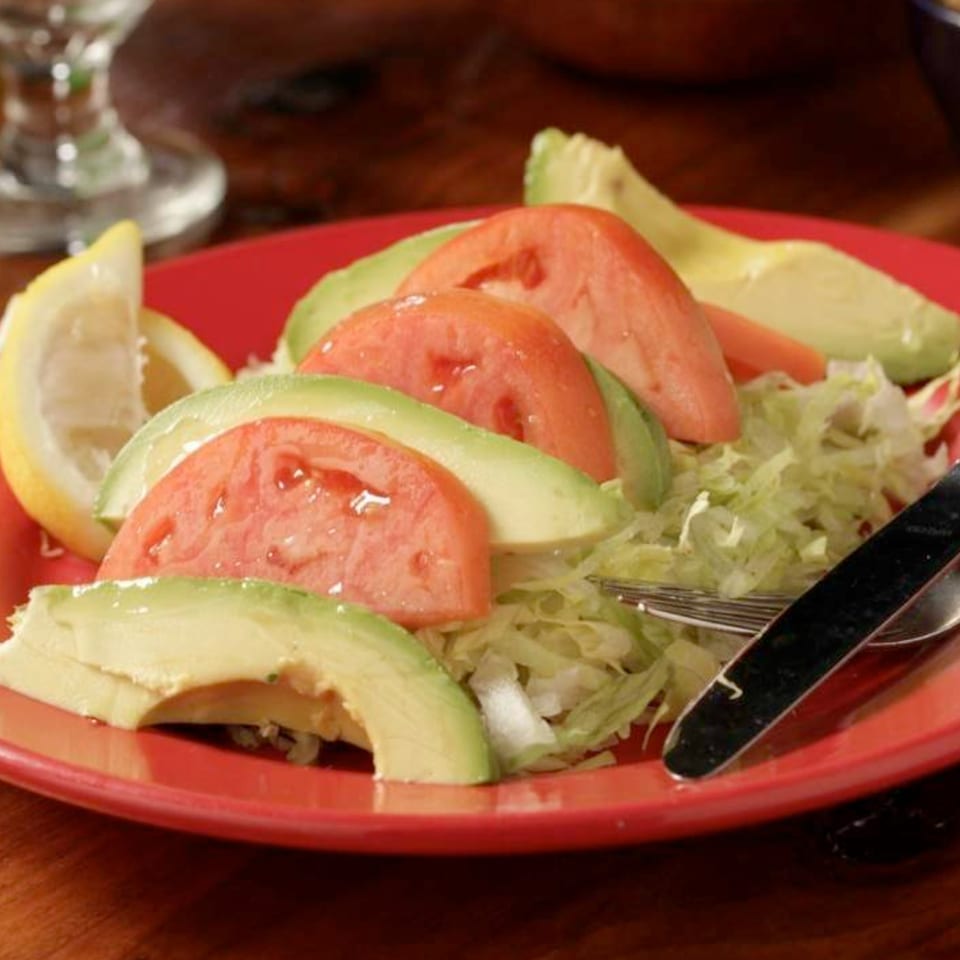 A red plate of shredded lettuce topped with sliced tomatoes, avocado, and lemon wedges.